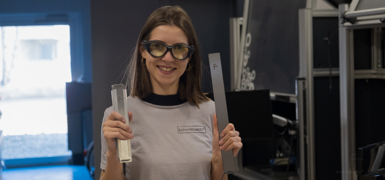 Gründerin und Entwicklerin Caroline Reiche hält in der Fertigung zwei Komponenten des modularen Schneidplatz-Systems SchnittKnecht® in den Händen: eine kompakt montierte Schneidebrett-Unterlage mit Silikonleisten sowie ein Edelstahl-Modul für die Erweiterung des Systems.
