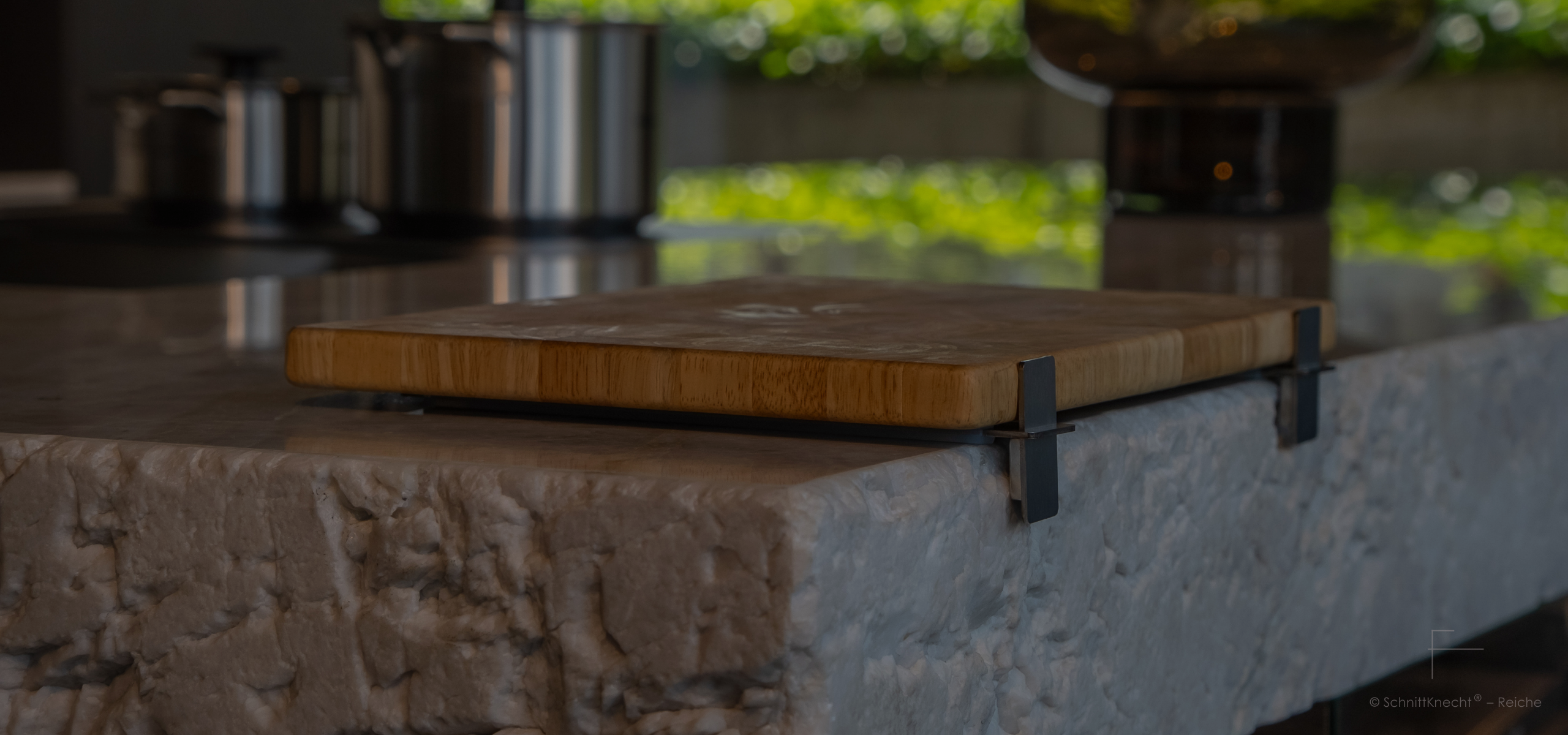 SchnittKnecht® Premium Schneidebrettunterlage aus Edelstahl und spülmaschinengeeignetem Silikon mit Schneidebrett in hochwertiger moderner Küche auf massiver Stein-Arbeitsplatte aus hellem Stein mit gebrochener Steinkante.