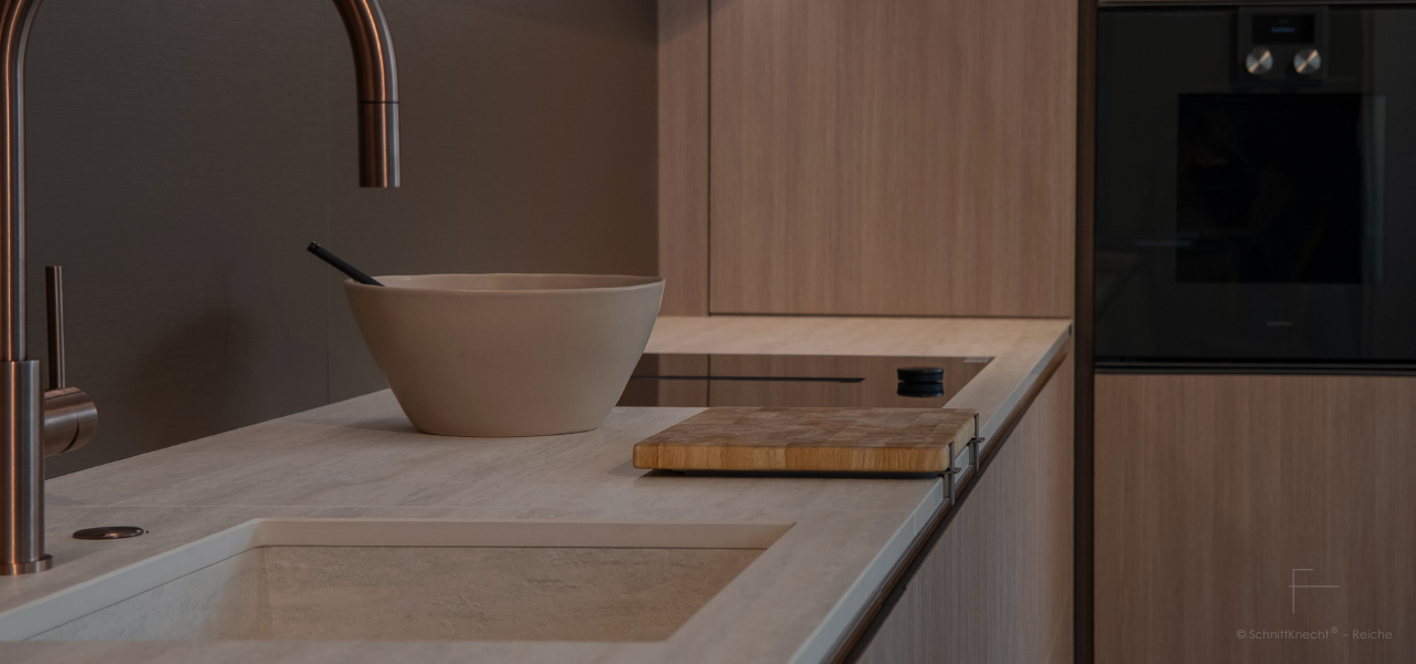 SchnittKnecht® Premium Schneidebrettunterlage aus Edelstahl und spülmaschinengeeignetem Silikon mit Schneidebrett in moderner heller Küche mit geriffelten Walnuss-Holzfronten, heller Steinplatte, Schüssel-Deko und eleganter Messing-Küchenarmatur.