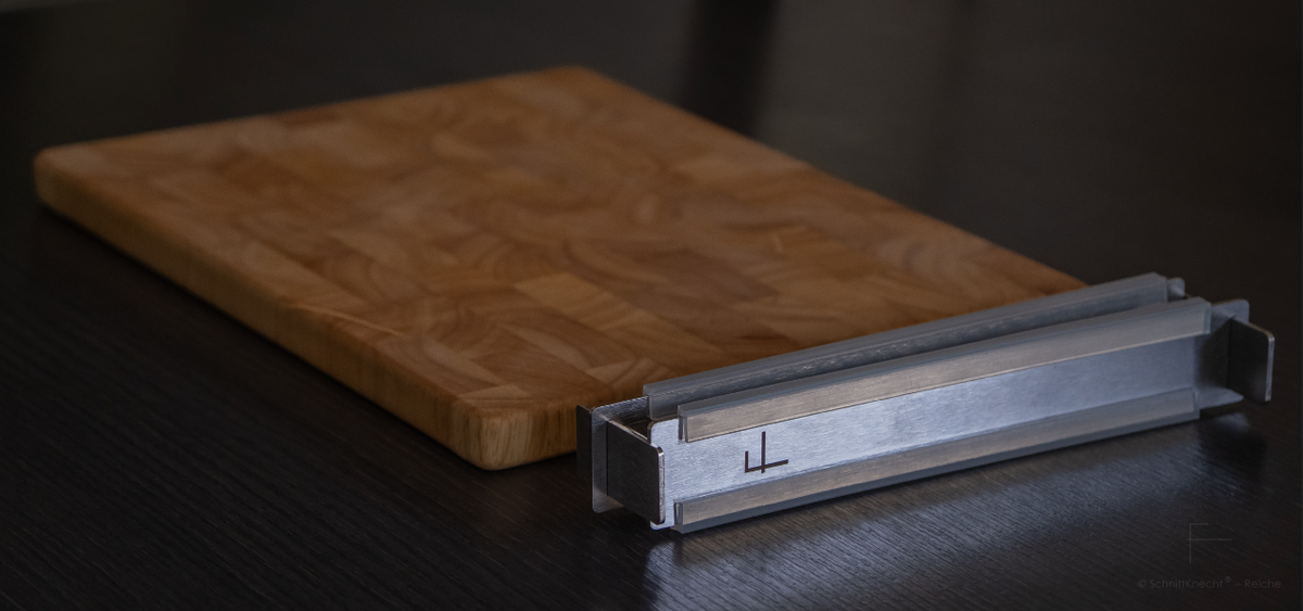 Kompakt zusammengebaute SchnittKnecht® Schneidebrett-Unterlage aus Edelstahl mit Silikonprofilen neben einem Holzschneidebrett auf dunkler Arbeitsplatte.