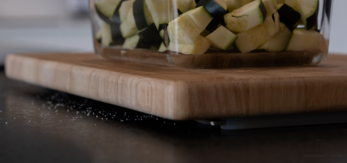 Nahaufnahme eines erhöhten Holzschneidebretts auf SchnittKnecht® mit Salzkörnern auf der dunklen Arbeitsplatte und sichtbarem Abstand unter dem Brett.