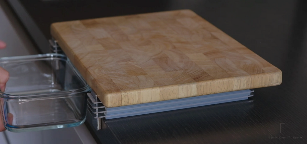 SchnittKnecht® Schneidebrett-Unterlage mit Holzschneidebrett, Ergonomie-Modulen und Behälter auf dunkler Küchenarbeitsplatte.
