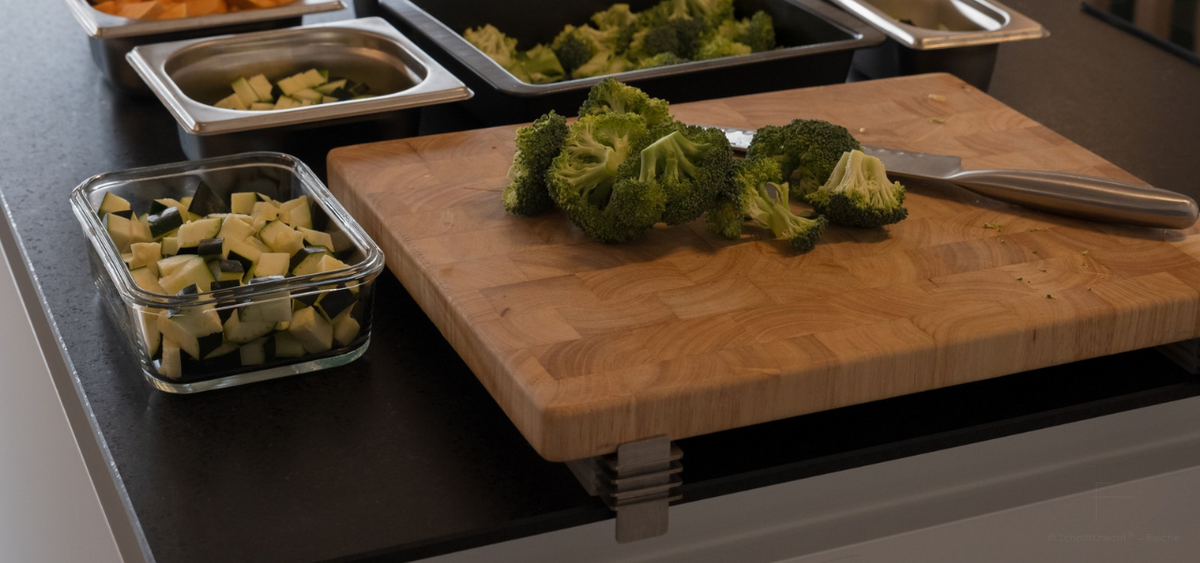 SchnittKnecht® Schneidebrett-Unterlage mit Ergonomie-Module und Holzschneidebrett, Brokkoli und Küchenmesser sowie einer Auflaufform, Glas- und Edelstahlbehältern mit vorbereitetem Gemüse.