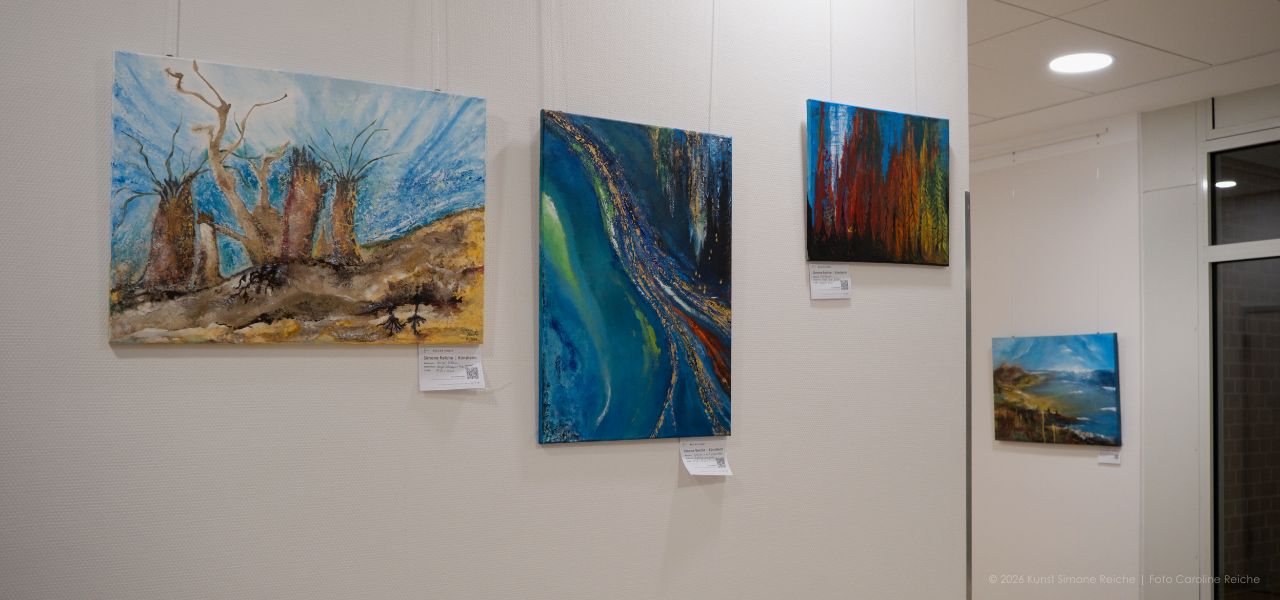 Flur des St. Anna Krankenhaus mit aufgehängten Gemälden von Künstlerin Simone Reiche im Rahmen der Ausstellung „Reiche Kunst“, darunter abstrakte Werke wie eine Einöde, eine Goldader, ein Waldbrand und zwei Angler am Meer.